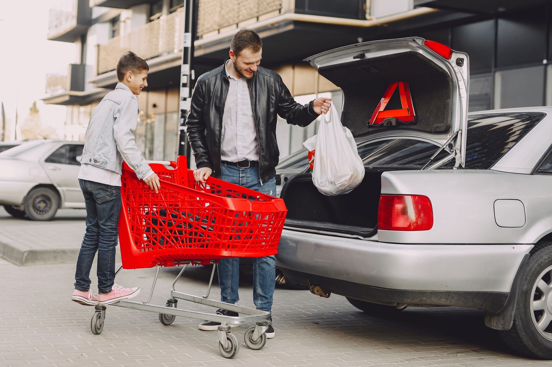 Happy parent and child loading grocery bags from cart into car trunk at curbside pickup.